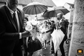 Une mariée, entourée de mains secourables, se dirige gracieusement vers l'église Saint-Pierre-et-Paul d'Ablis, en France, lors d'une forte tempête de pluie, capturée dans une image intemporelle en noir et blanc.